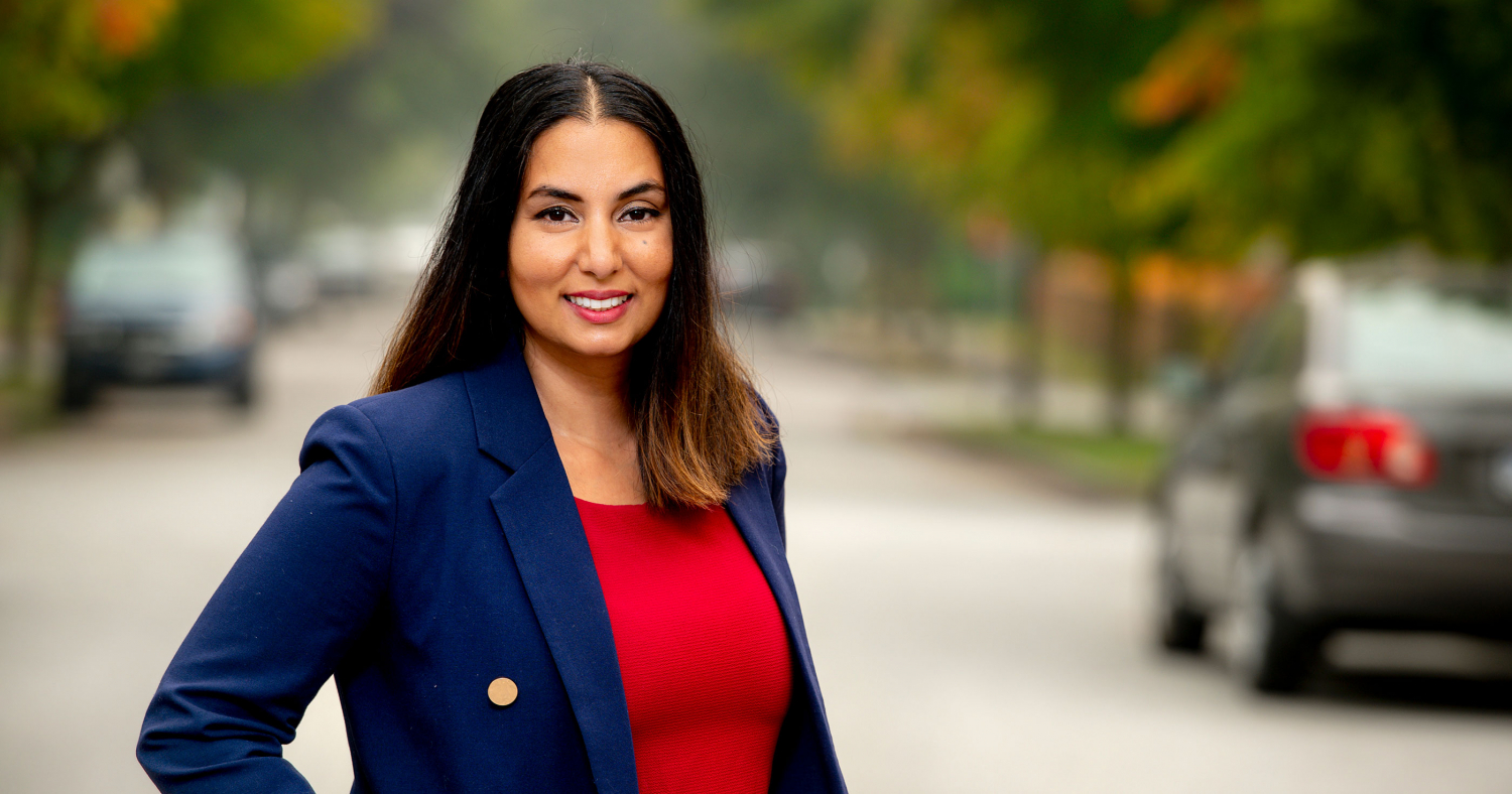 A smiling, confident woman of colour in a red shirt and dark blue blazer standing the street